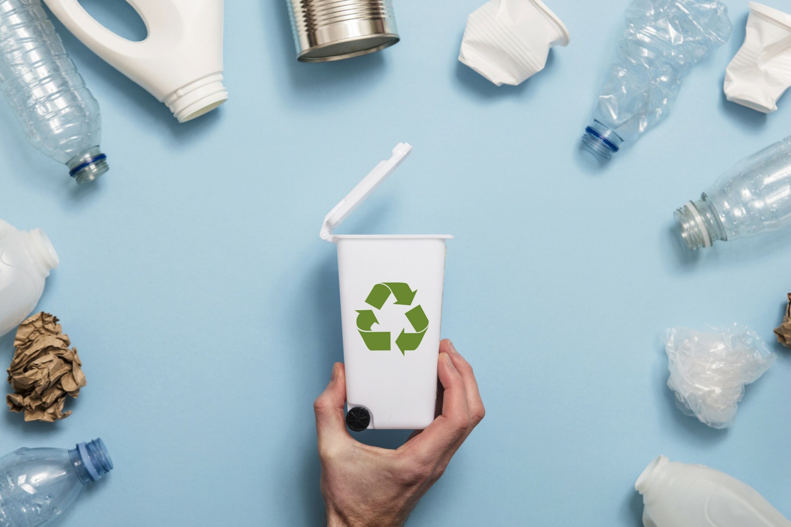  A hand holding a white recycling bin with a green recycling symbol surrounded by plastic bottles, cans, and other recyclable materials.